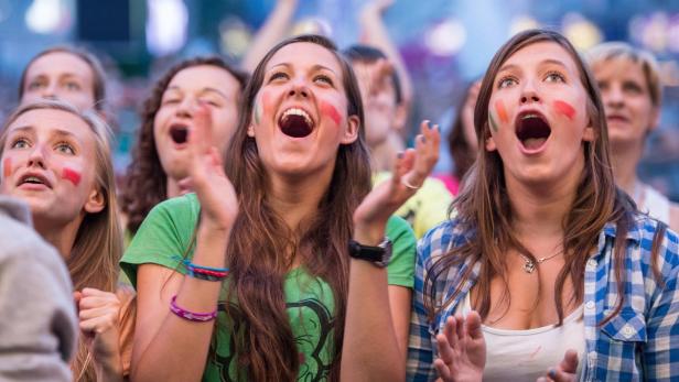 Eine Gruppe junger Frauen jubelt mit aufgemalten Flaggen auf ihren Wangen.