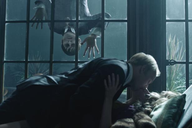 Ein Paar küsst sich auf einem Sofa, während Wednesday Addams durch ein Fenster schaut.