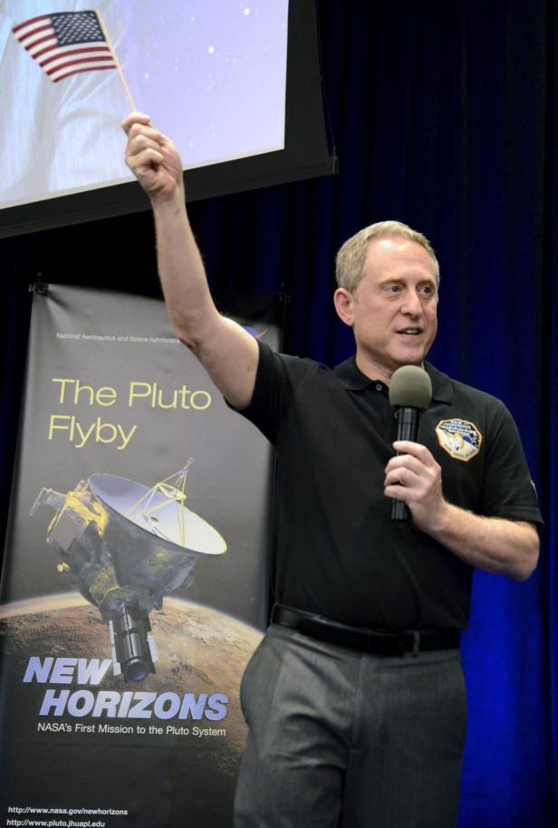 Ein Mann spricht am Mikrofon und hält eine amerikanische Flagge hoch, im Hintergrund ein „New Horizons“-Poster.