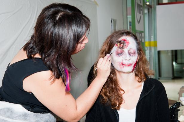 Eine Frau schminkt eine andere Frau als Zombie mit einer Wunde auf der Stirn.
