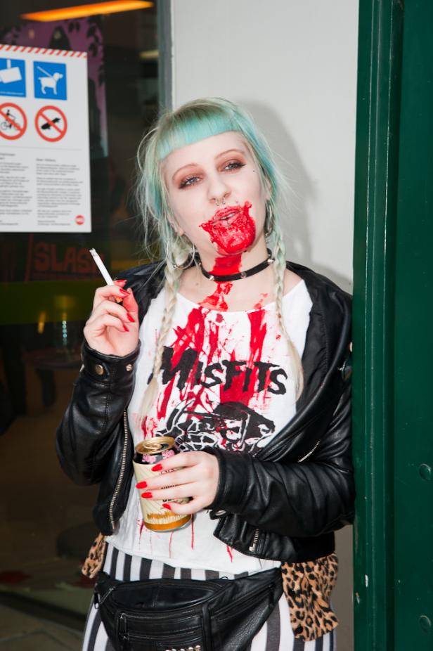 Eine Frau mit blauen Haaren, „Misfits“-T-Shirt und Kunstblut trinkt Bier und raucht eine Zigarette.