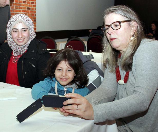 Eine Frau mit Brille zeigt einem Kind und einer jungen Frau mit Kopftuch etwas auf einem Smartphone.