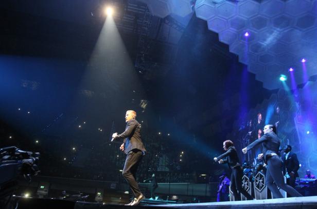 Robbie Williams tritt mit Tänzern auf einer Bühne vor Publikum auf.