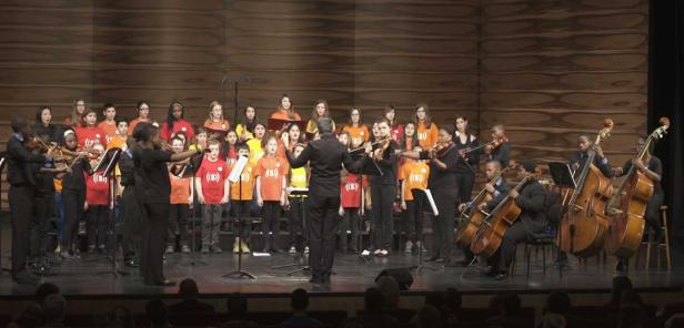 Ein Kinderchor und ein Orchester treten auf einer Bühne auf.