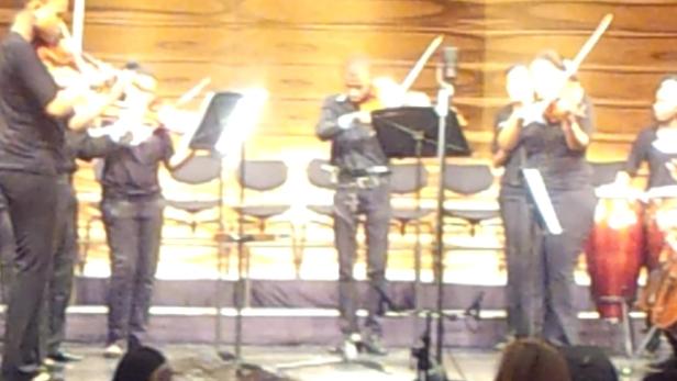 Ein Orchester spielt auf einer Bühne mit Geigen und Perkussion.