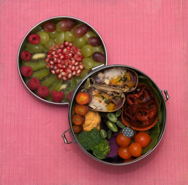 Eine Lunchbox mit Wraps, Gemüse und eine Schale mit Obst auf einem rosa Tuch.