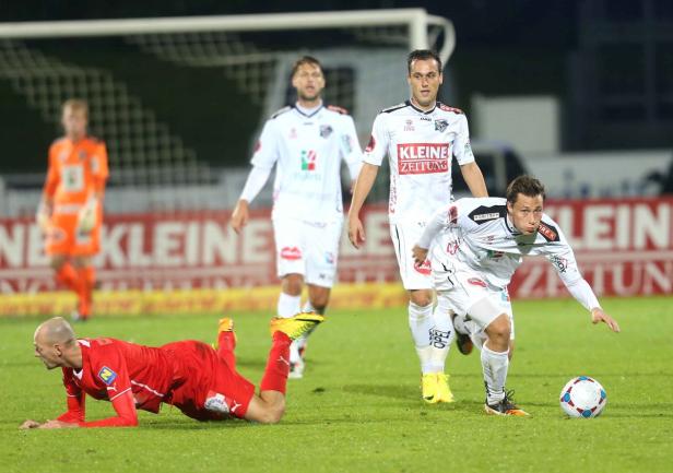 Ein Fußballspieler in Rot liegt am Boden, während ein Spieler in Weiß den Ball kontrolliert.