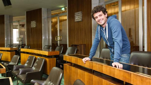 Ein lächelnder Mann in einem blauen Hoodie lehnt an einem Tisch in einem Konferenzraum.