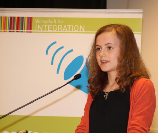 Eine junge Frau spricht am Rednerpult vor einem Banner „Wirtschaft für Integration“.