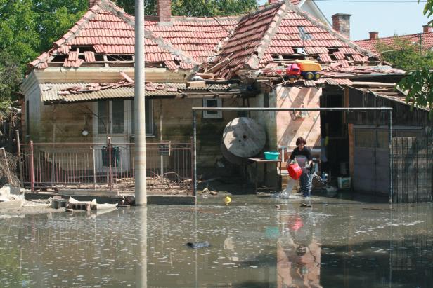 Eine Frau schöpft Wasser vor einem beschädigten Haus, auf dessen Dach Spielzeugautos stehen.