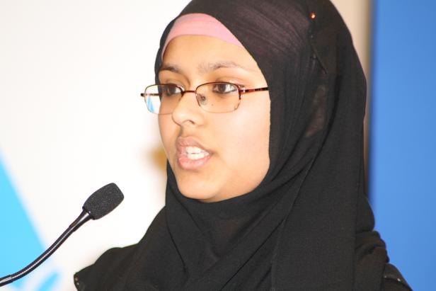 Eine junge Frau mit Hijab und Brille spricht in ein Mikrofon.
