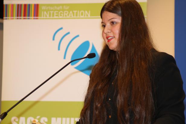 Eine Frau spricht bei einer Veranstaltung vor einem Banner „Wirtschaft für Integration“.