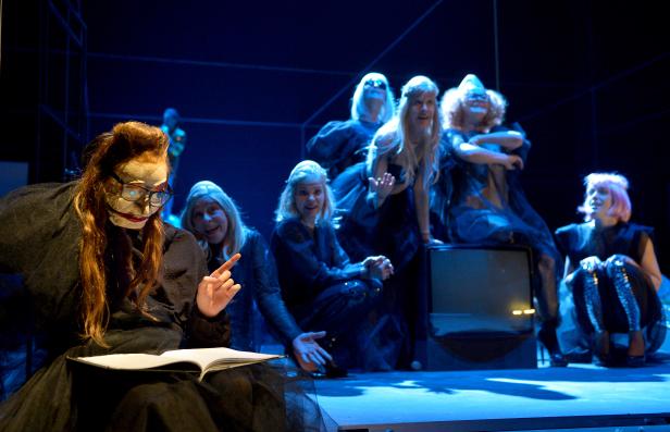 Eine Theaterszene mit einer Gruppe von Schauspielerinnen in Kostümen und Masken auf einer Bühne.