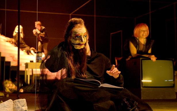 Eine Person mit einer Maske sitzt mit einem Buch auf einer Bühne.
