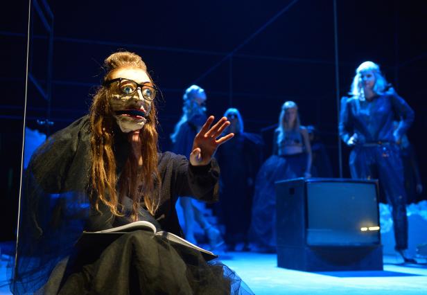 Eine Person mit einer grotesken Maske liest auf einer Bühne ein Buch.