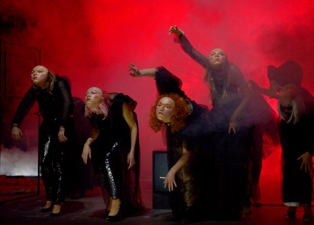 Eine Gruppe von Performerinnen in schwarzen Kostümen und Masken auf einer Bühne mit rotem Licht.
