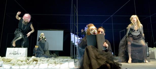 Eine Theaterszene mit mehreren Frauen in schwarzen Kleidern und einem Mann mit einer Maske, die ein Buch liest.