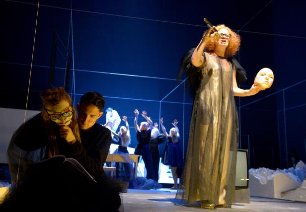 Eine Frau im goldenen Kleid hält eine Maske auf einer Theaterbühne.