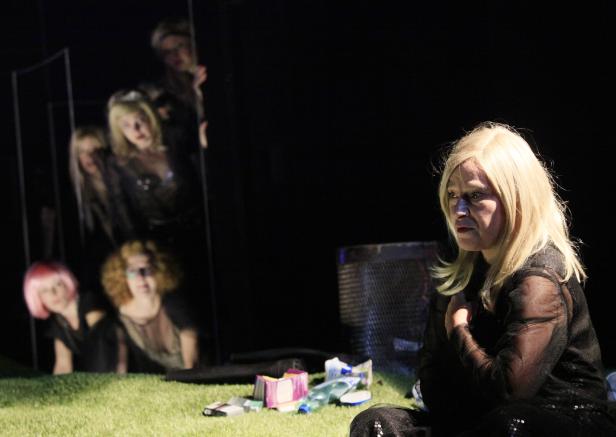 Eine Frau mit blonden Haaren sitzt auf einer Wiese, während andere Frauen durch einen Rahmen im Hintergrund schauen.