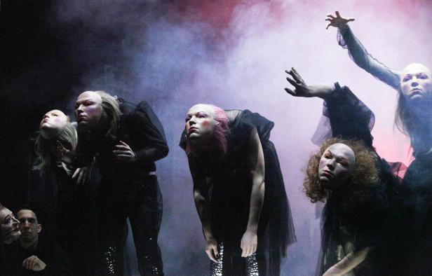 Eine Gruppe von Performer*innen mit blass geschminkten Gesichtern auf einer Bühne.