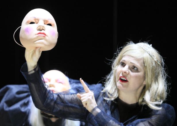 Eine Frau mit Krone hält eine weiße Maske in der Hand.