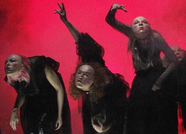 Mehrere Performer in dunklen Kostümen und theatralischem Make-up vor einem roten Hintergrund.