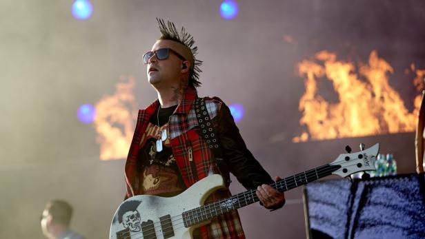 Ein Musiker mit Irokesenschnitt spielt Bassgitarre vor einem Hintergrund aus Flammen.