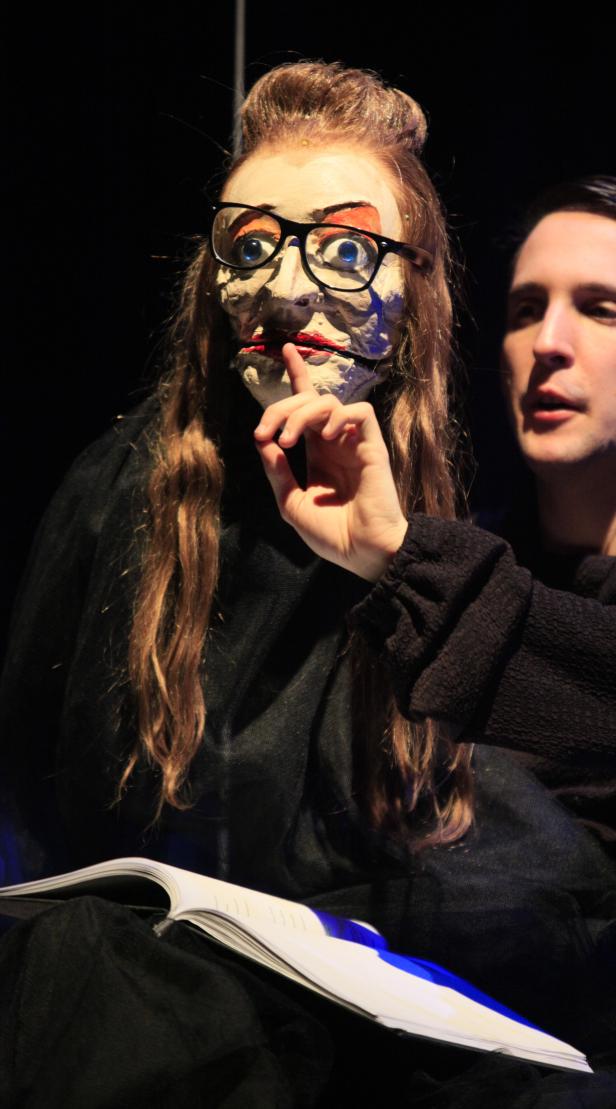 Ein Mann interagiert mit einer Puppe mit übergroßer Maske und Brille, möglicherweise im Theater.