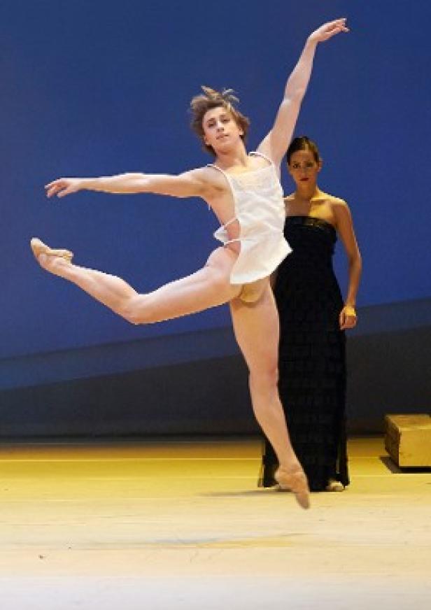 Ein männlicher Balletttänzer springt vor einer Frau in einem schwarzen Kleid auf der Bühne.