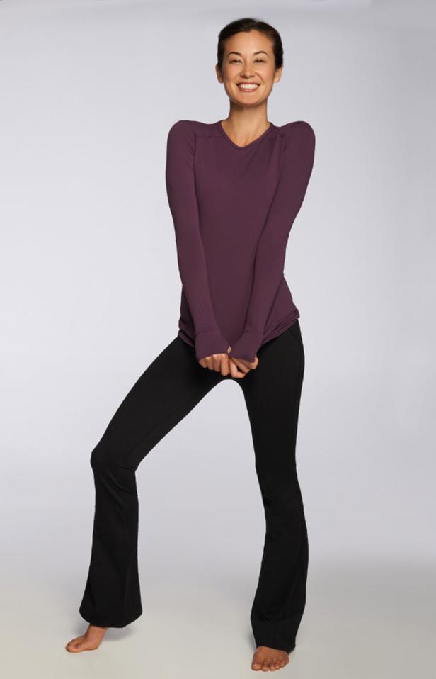 Eine lächelnde Frau in Sportkleidung posiert barfuß vor einem hellen Hintergrund.