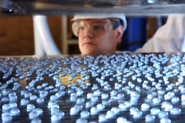 Ein Arbeiter mit Schutzbrille und Helm betrachtet kleine, halbkugelförmige Pellets auf einer Metalloberfläche.