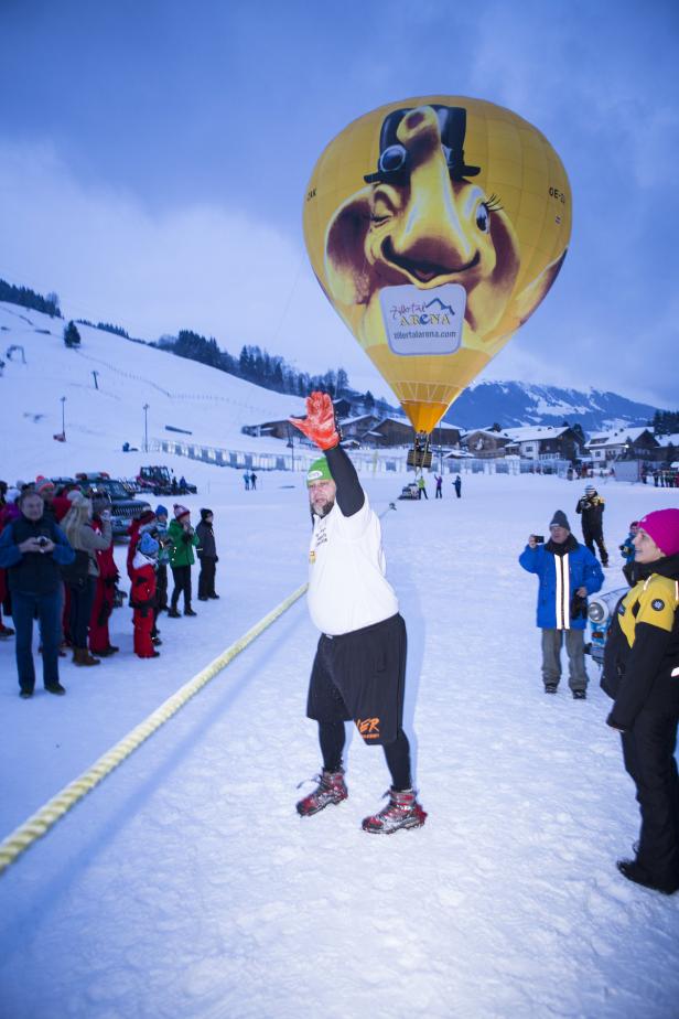 Ein Mann winkt vor einem gelben Heißluftballon mit einem Gesicht darauf im Schnee.