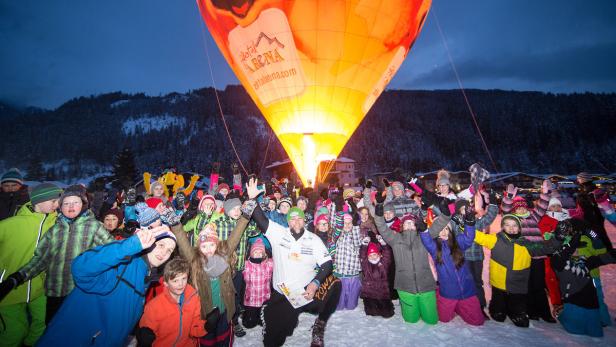 Eine Gruppe Kinder jubelt vor einem beleuchteten Heißluftballon im Schnee.