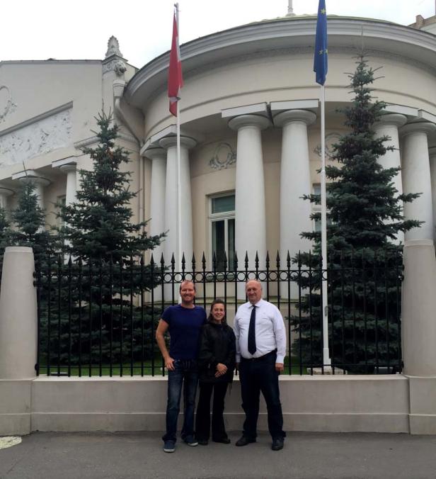 Drei Personen stehen vor einem Gebäude mit Säulen, einer lettischen und einer EU-Flagge.