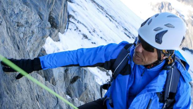 Ein Bergsteiger mit Helm und Sonnenbrille klettert eine steile Felswand hinauf.