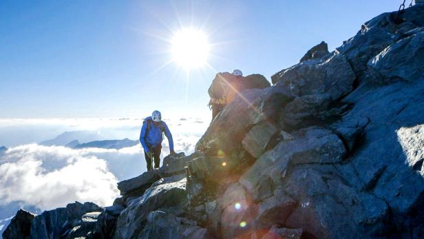 Zwei Bergsteiger erklimmen einen felsigen Gipfel bei strahlendem Sonnenschein.