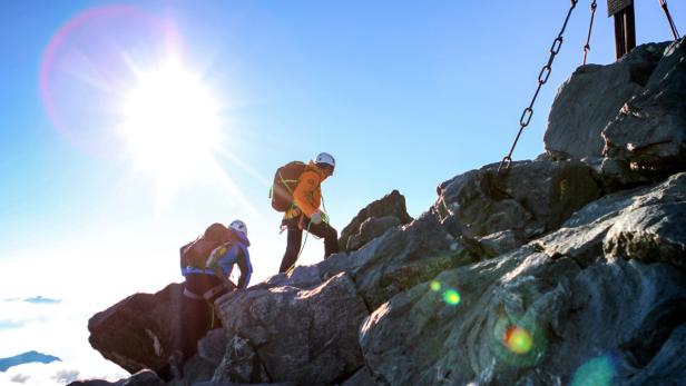 Zwei Bergsteiger erklimmen einen felsigen Gipfel bei strahlendem Sonnenschein.
