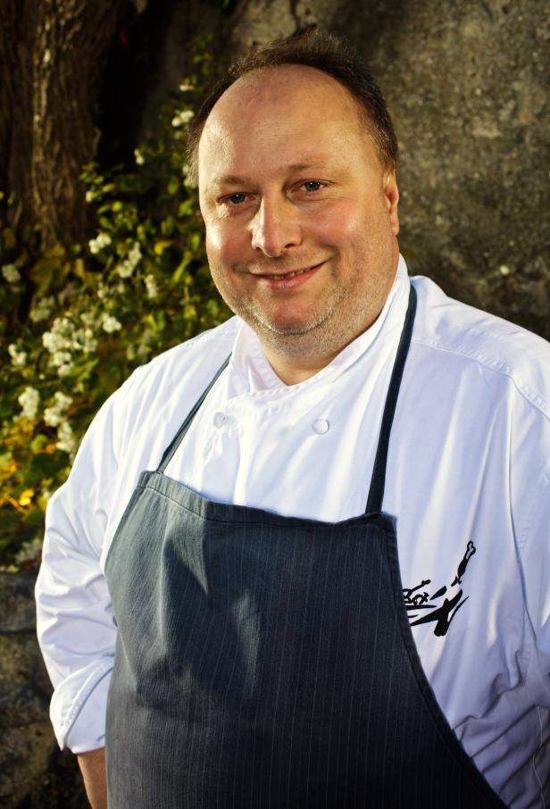 Ein lächelnder Koch in Kochjacke und Schürze steht im Freien.