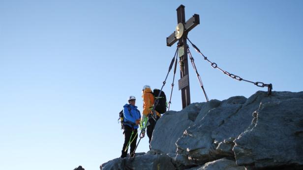 Zwei Bergsteiger stehen neben einem Gipfelkreuz auf einem felsigen Gipfel.