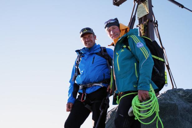 Zwei Bergsteiger stehen am Gipfelkreuz und blicken in die Kamera.