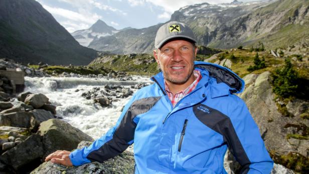 Ein Mann in Berglandschaft mit blau-schwarzer Jacke und Kappe vor einem reißenden Fluss.