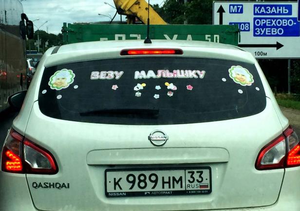 Ein weißer Nissan Qashqai mit Aufklebern auf der Heckscheibe, darunter „ВЕЗУ МАЛЫШКУ“.