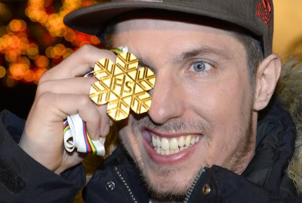 Ein Mann hält eine goldene FIS-Medaille vor sein Auge und lächelt.