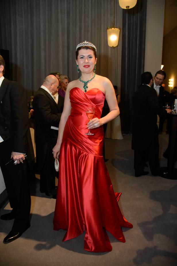 Eine Frau in einem roten Abendkleid, mit Tiara und Schmuck, hält ein Glas Sekt.