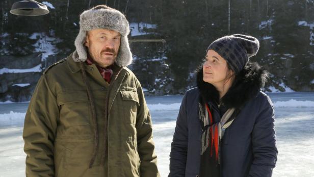 Ein Mann mit Fellmütze und eine Frau mit Mütze stehen im Winter vor einer Eisfläche.