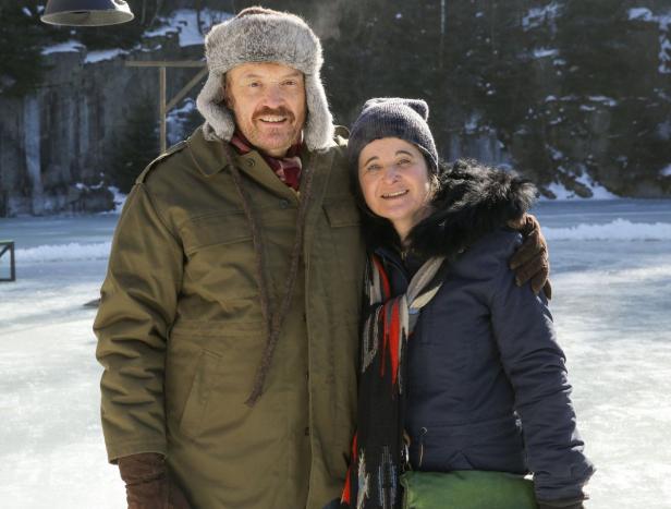Ein Mann mit Fellmütze und eine Frau mit Mütze stehen im Winter vor einer Eisfläche.