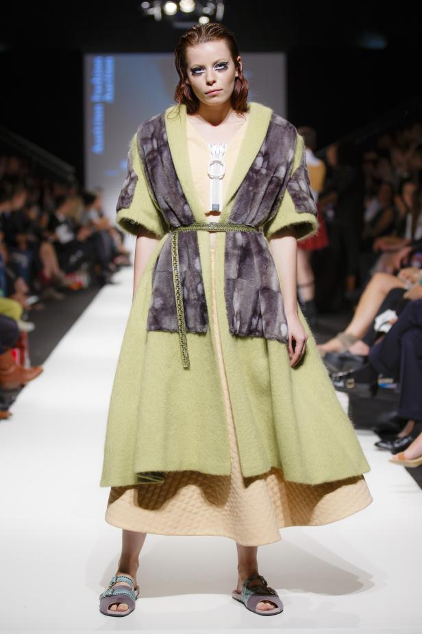 Eine Frau präsentiert ein grünes Kleid mit Fellbesatz auf einem Laufsteg.