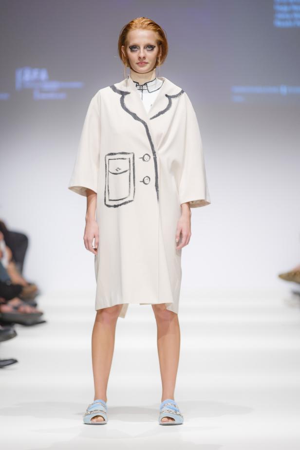 Eine Frau präsentiert einen hellen Mantel mit aufgemalten Details auf einem Laufsteg.