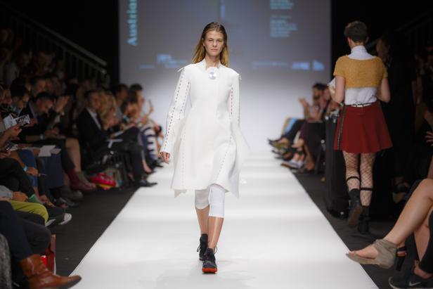 Eine Frau präsentiert ein weißes Kleidungsstück auf einem Laufsteg vor Publikum.