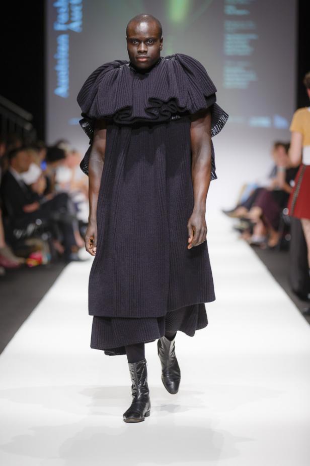 Ein dunkelhäutiger Mann präsentiert ein dunkles, voluminöses Strickkleid auf dem Laufsteg.
